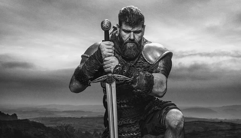 Bearded man in Viking-style armor holding a large sword against a cloudy landscape.