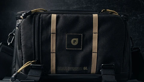 Black camera bag with tan stripes and brass hardware against dark background.