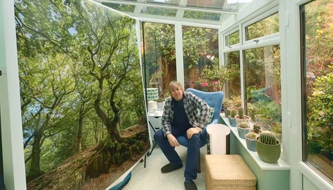 Man in plaid shirt crouching in sunlit conservatory with forest mural and potted plants.