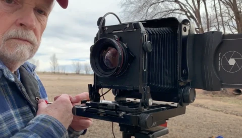 Photographer holding a large format camera with bellows and ground glass in an open field.
