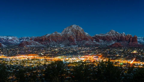 Snow-dusted red rock formations rise above a town illuminated by warm lights at dusk.