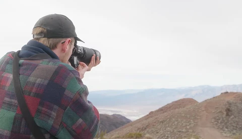 Photographer with camera raised to face, wearing plaid shirt and cap, standing on hillside overlooking desert valley.