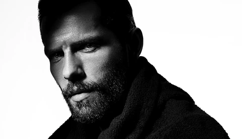 Bearded man with dark hair in black and white portrait photography, shot against white background with dramatic side lighting.