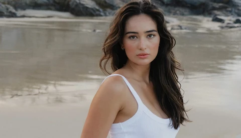 Young woman in white tank top posing at beach with rocky outcrops behind her.