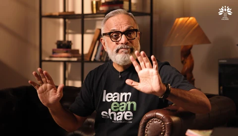 Man with gray beard and glasses wearing black shirt gesturing while speaking in warm interior setting.