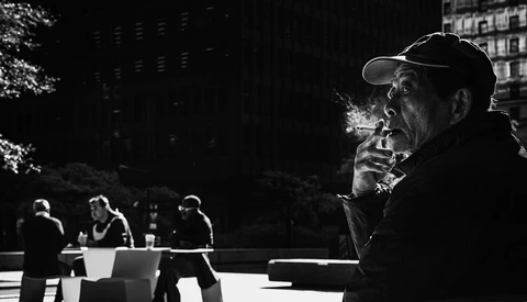 Man in baseball cap smoking cigarette in urban plaza with other figures blurred in background.