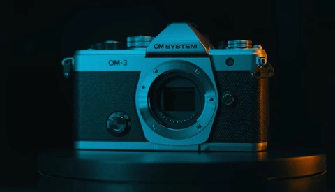 OM System OM-3 rangefinder camera with cyan and orange gel lighting against dark background.