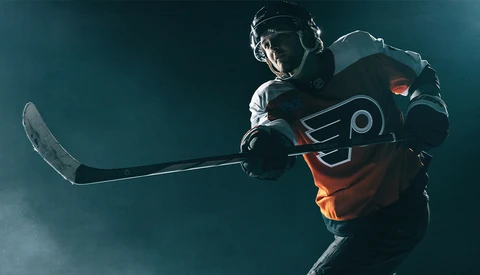 Hockey player in orange and black jersey holding stick against dark blue background with atmospheric lighting.