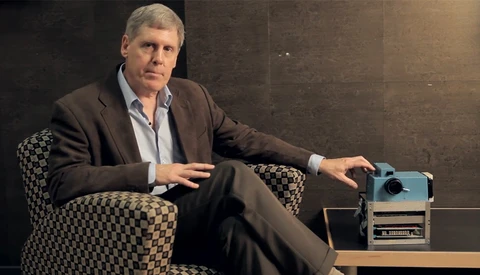 Man in business casual attire seated in patterned chair next to vintage projector.