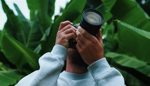 Photographer holding a telephoto lens up to frame a shot among large green leaves.
