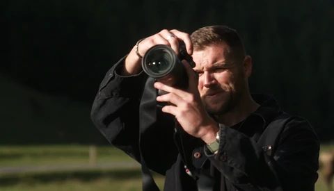 Photographer in black jacket holding camera with telephoto lens to his eye, focused on subject with concentrated expression.