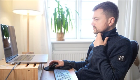Man at desk reviewing laptop screen while working with keyboard and mouse in bright home office.