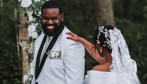 Groom in light blue suit smiling at bride during outdoor wedding ceremony.