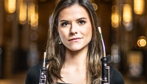 Portrait of a young woman holding oboes against a warm, blurred interior background.