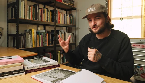 Man in black sweatshirt and cap gestures while discussing photography work at wooden desk surrounded by books and shelves.