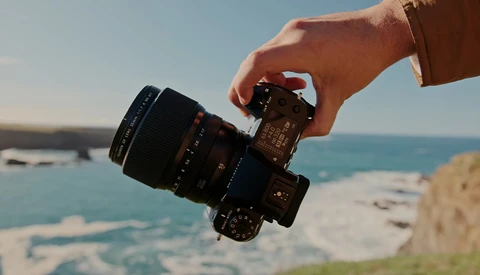 Hand holding a mirrorless camera with attached lens, positioned against a coastal seascape with turquoise water and rocky cliffs.