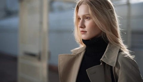 Woman in beige coat and black turtleneck gazing to the side in natural window light.