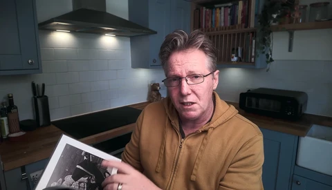 Man in glasses and tan jacket holding a black and white photograph in a modern kitchen.