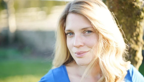 Woman with blonde hair smiling at camera in natural outdoor lighting.