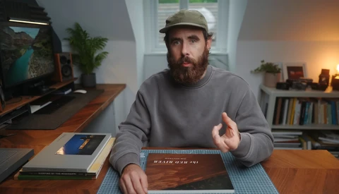Bearded man wearing a gray sweatshirt and cap gestures while discussing photography books at a wooden desk.