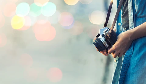 Photographer in denim shirt holding a vintage rangefinder camera with soft-focus bokeh lights in background.