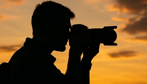 Silhouetted photographer holding a DSLR camera against a golden sunset sky.