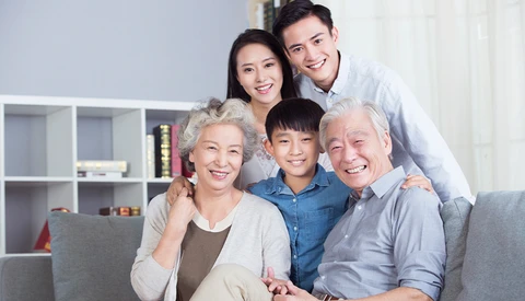 Multi-generational Asian family of five smiling together on a gray couch in a modern living room.