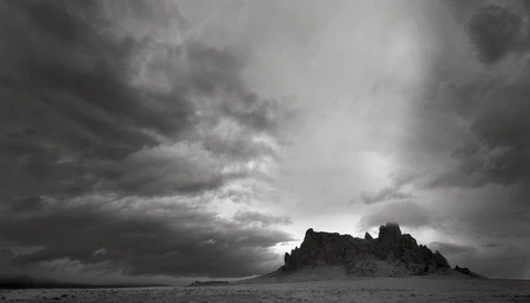 Silhouetted rocky pinnacles rise from a flat desert plain beneath a dramatic storm sky.