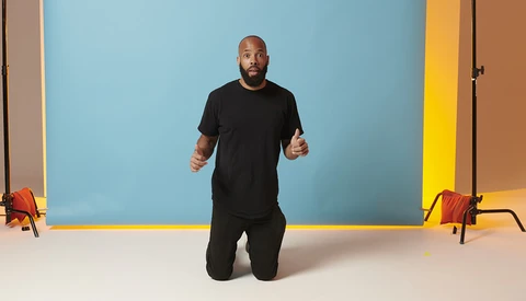 Man in black clothing kneeling on white floor against blue backdrop with yellow trim.