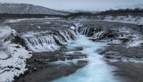 Frozen waterfall cascading through snow-covered landscape with turquoise glacial water flowing between dark rocks.
