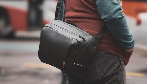 Person wearing a black camera backpack with shoulder strap over casual clothing in an urban street setting.