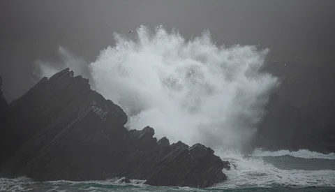 Massive ocean wave crashes against dark rocky coastline, creating explosive white spray and mist.