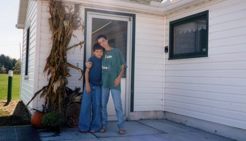 Two people standing in a doorway of a white residential building with autumn decorations nearby.