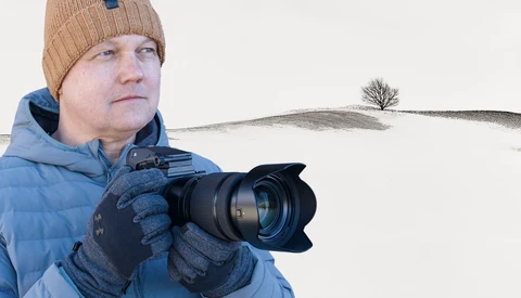 Photographer in winter gear holding a DSLR camera with hood lens in snowy landscape.