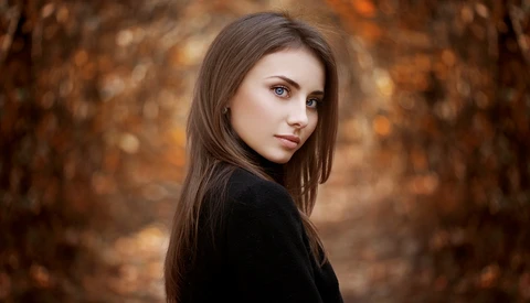 Young woman with long dark hair gazing over her shoulder against a blurred autumn forest background.