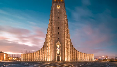 Illuminated modern church with soaring triangular architecture at dusk, featuring vertical ribbed columns and a glowing cross window.