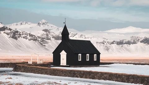 Black wooden church with white door and cross standing alone in snow-covered landscape beneath snow-capped mountains.