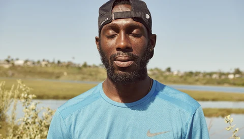 Man wearing light blue Nike t-shirt and dark cap standing outdoors with wetland landscape behind him.