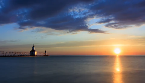 Lighthouse silhouetted against golden sunset over calm water with dramatic blue clouds.