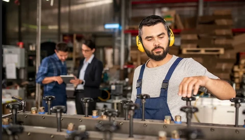 Factory worker in yellow hearing protection adjusts machinery while colleagues confer in the background.