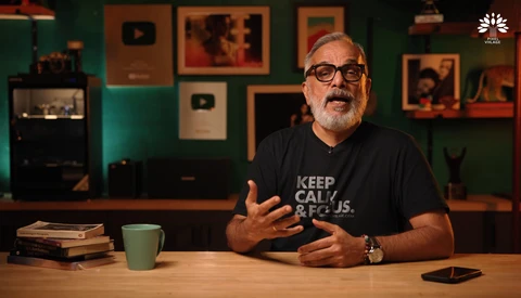Man wearing glasses and black t-shirt gestures while speaking at wooden desk in studio setting.