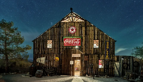Weathered wooden general store at night beneath a starlit sky, illuminated by warm interior light and vintage neon signage.