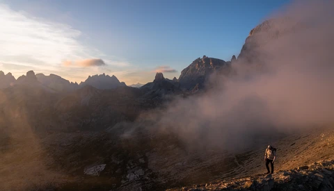 Hiker standing on a rocky slope with dramatic mountain peaks and misty valleys at golden hour.