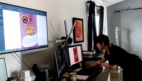 Photographer working at desk with multiple monitors displaying food photography and editing software.