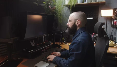Bearded man working at a computer desk in a dark studio space surrounded by plants and equipment.