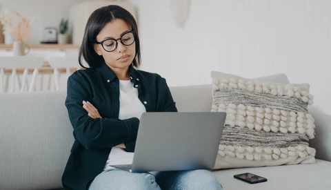 Woman wearing glasses and dark blazer sits on gray couch working on laptop with arms crossed.
