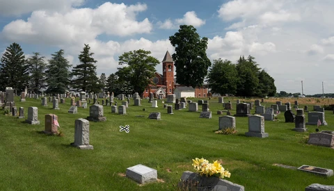 Cemetery landscape with gravestones, green grass, flowering arrangement, and brick church with steeple in background under partly cloudy sky.