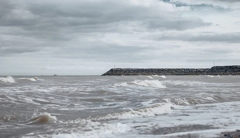 Rough ocean waves breaking on a beach under overcast sky with a rocky breakwater visible in the distance.