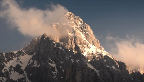 Snow-covered mountain peak with golden sunlit summit and dramatic clouds swirling around the ridges.