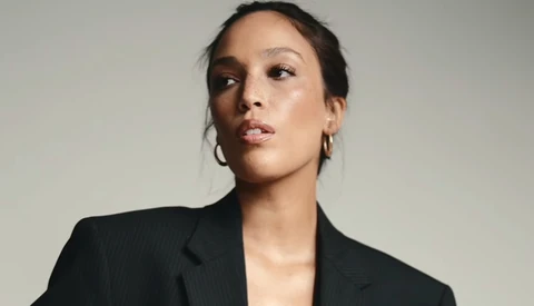 Woman in black pinstriped blazer against neutral background, looking upward with composed expression.